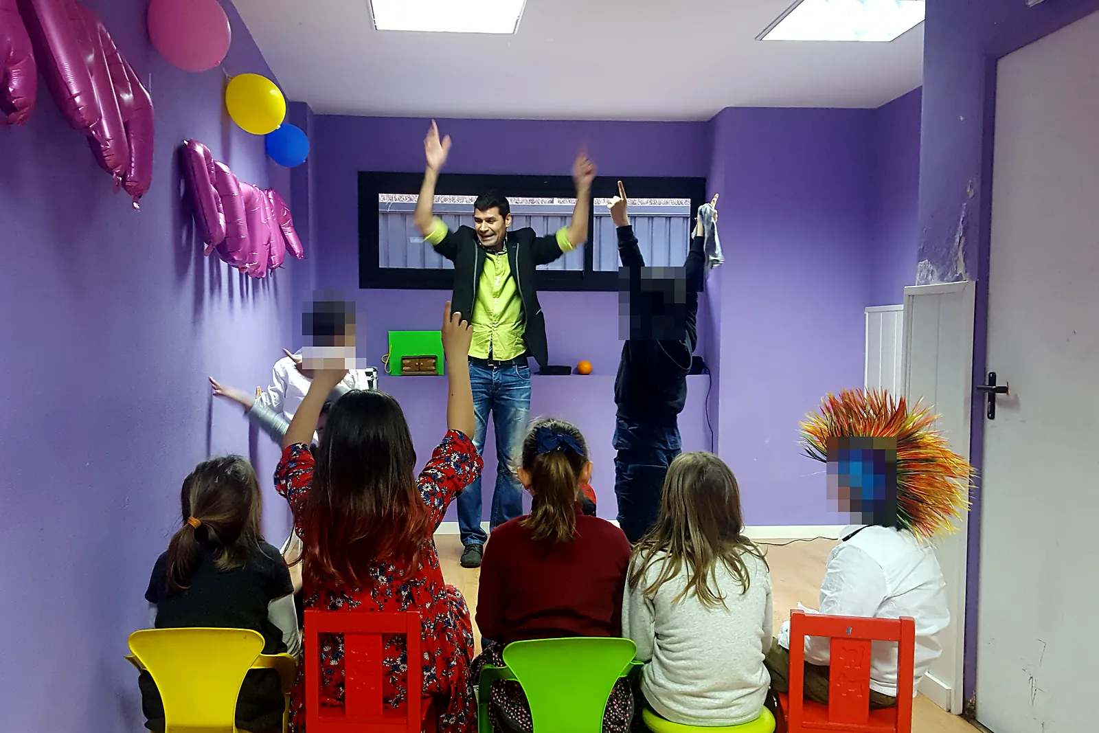 Niños sentados en sillas de colores levantando la mano mientras el mago actúa con dos voluntarios